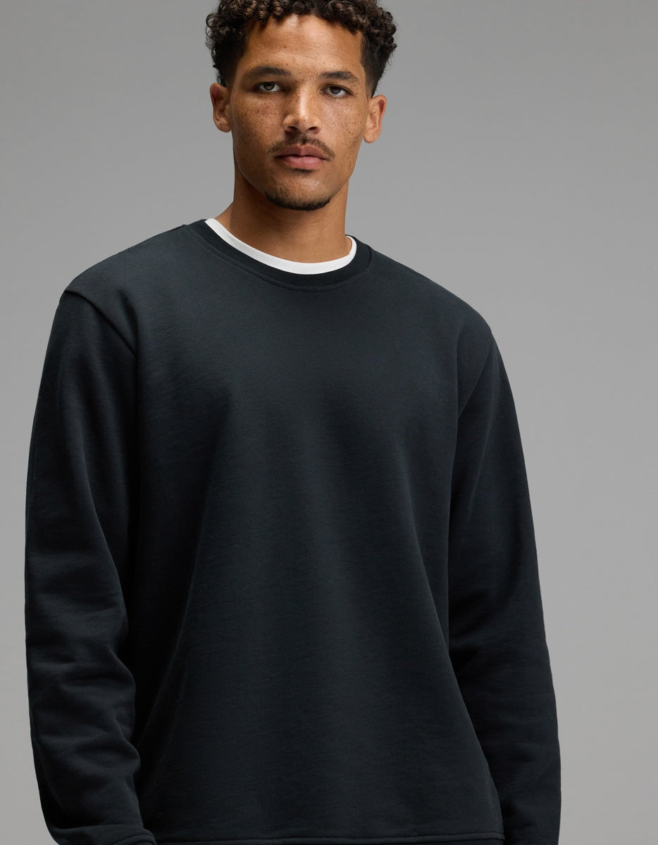 A man wearing the Men’s Dylan Crewneck in Jet Black by Cozy Earth over a white shirt stands against a plain gray background, looking toward the camera with a neutral expression.|Color:Jet Black