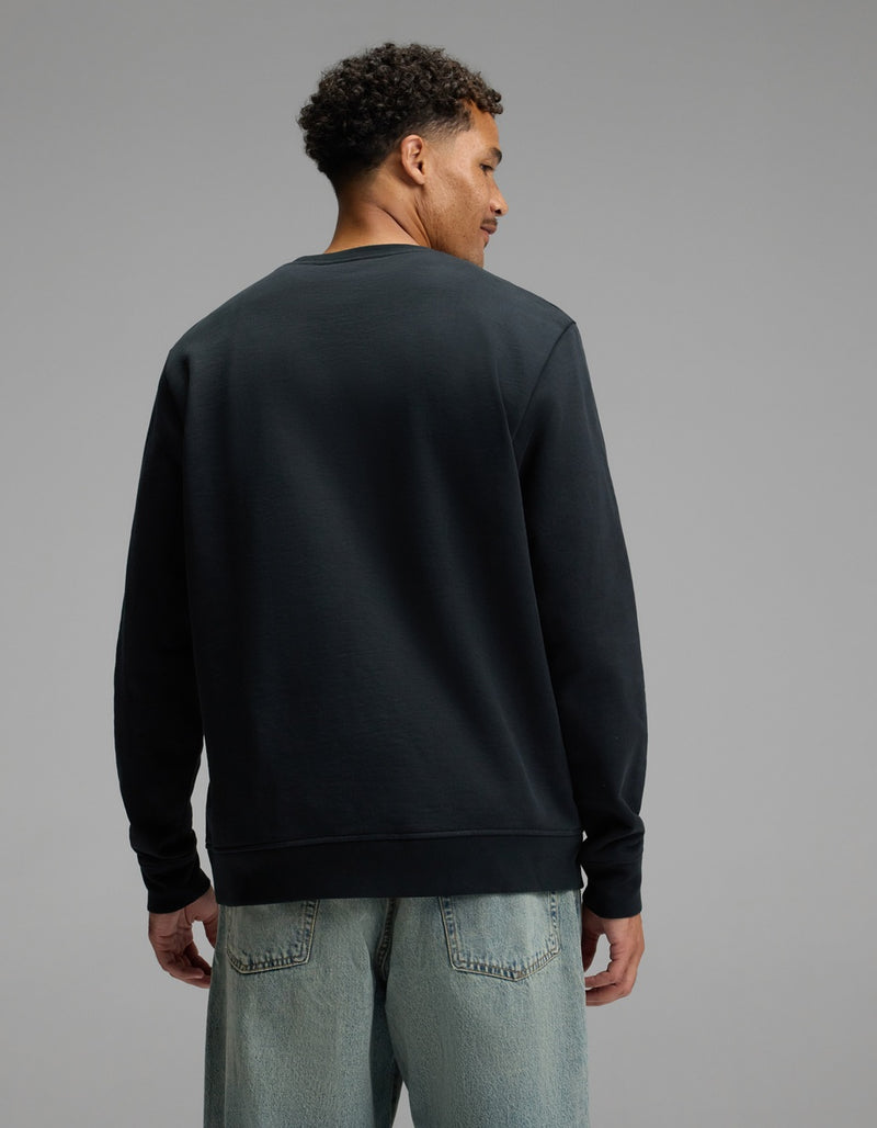 A man stands facing away, wearing the Cozy Earth Men’s Dylan Crewneck in black and light blue jeans, against a plain gray background.