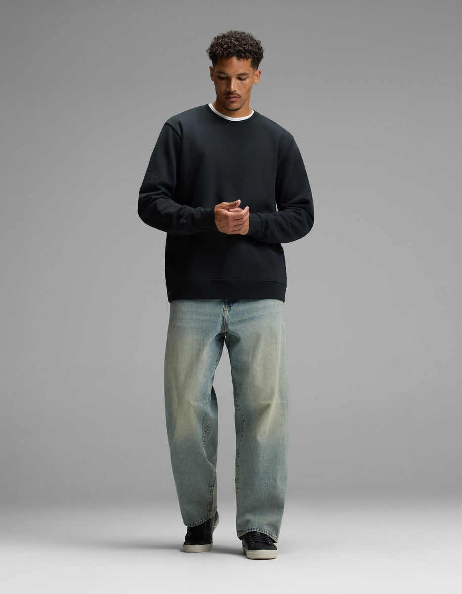 A man stands against a plain gray background, wearing the Cozy Earth Men’s Dylan Crewneck, paired with light blue relaxed-fit jeans and black sneakers. He looks down with his hands clasped together in front of him.