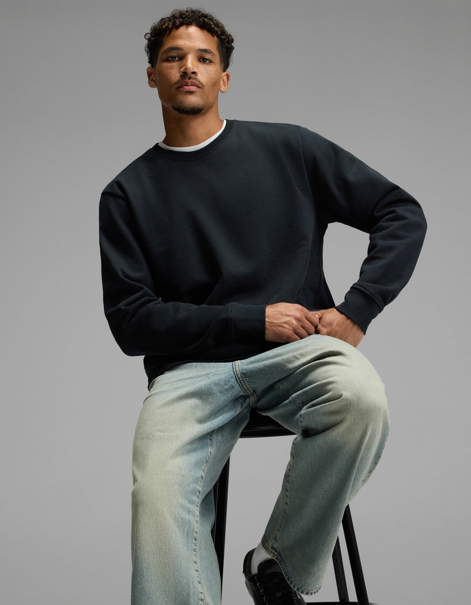 A man with short curly hair sits on a stool, wearing the Cozy Earth Men’s Dylan Crewneck, light blue jeans, and black sneakers, posing against a plain gray background.|Color:Jet Black