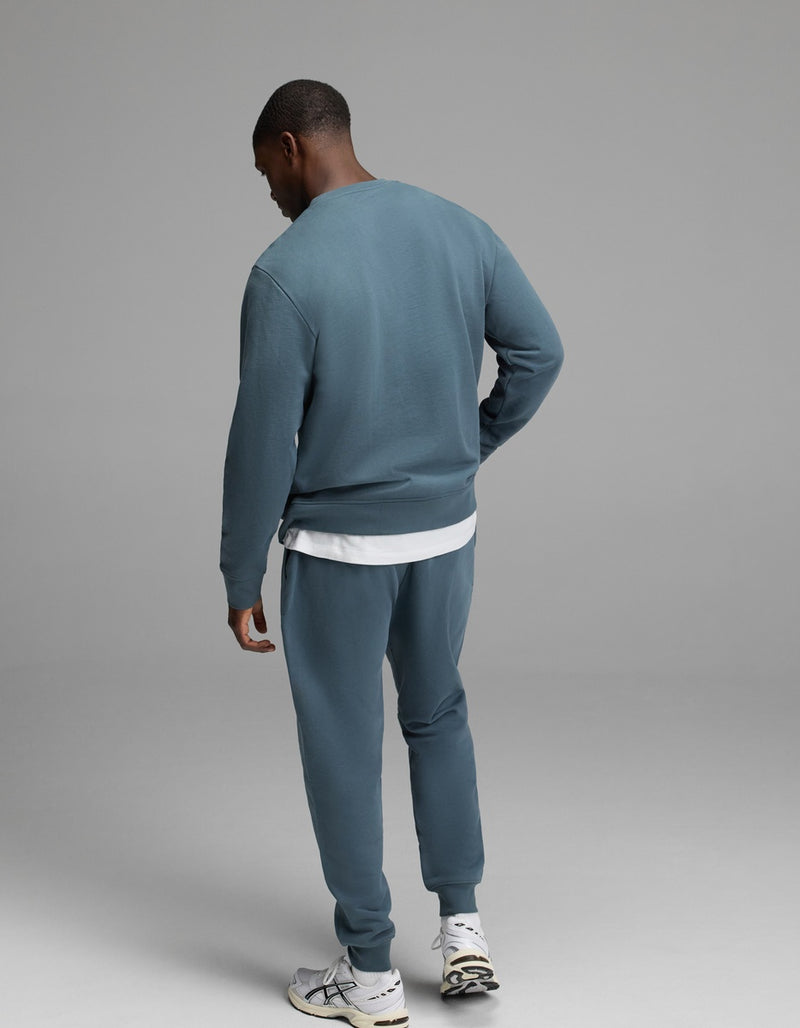 A person stands facing away against a plain gray background, wearing the Cozy Earth Men’s Dylan Crewneck & Jogger Set in blue-green over a white T-shirt and paired with white sneakers.