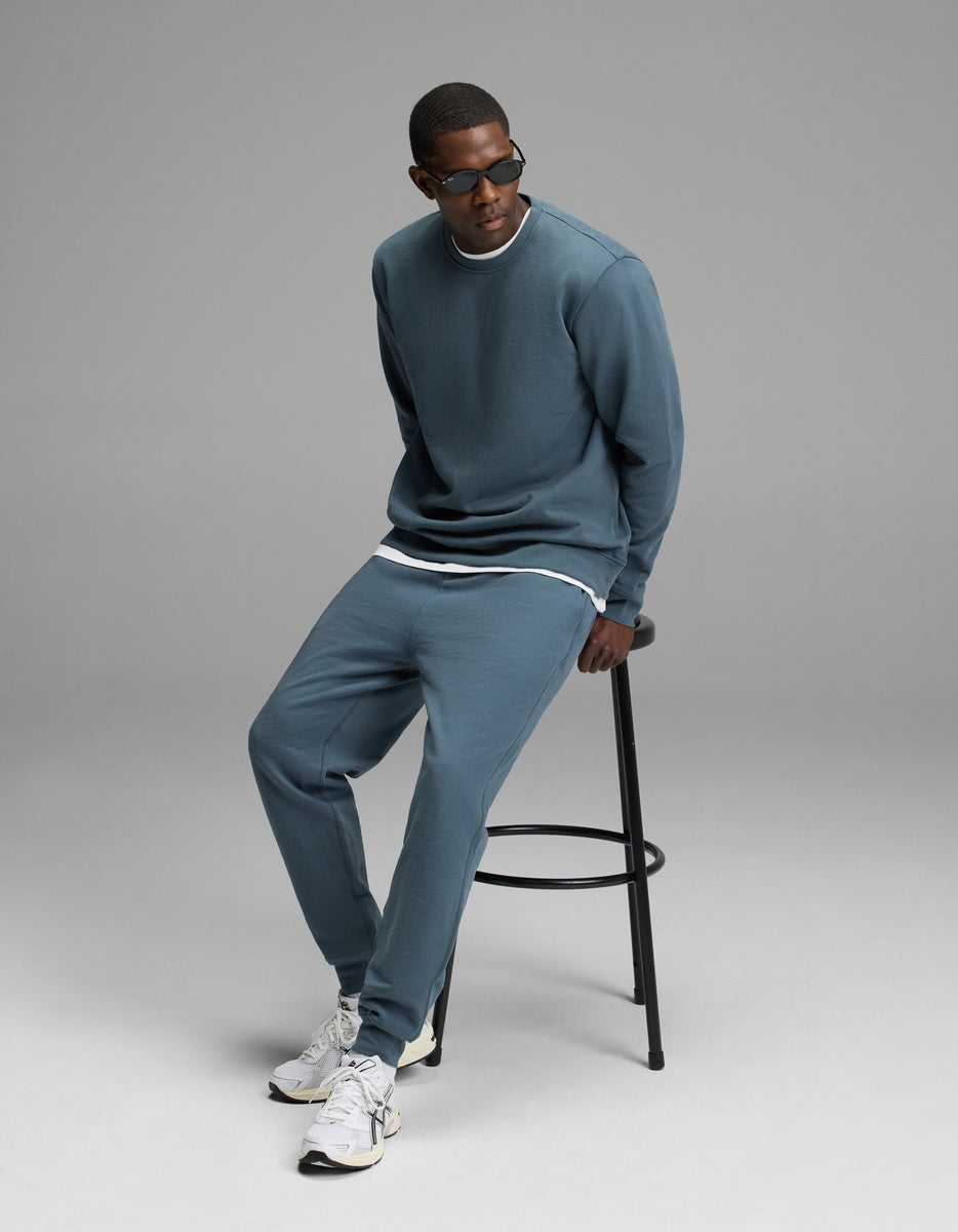 A person wearing the Men’s Dylan Crewneck & Jogger Set by Cozy Earth sits on a black stool against a plain gray background. They are also wearing sunglasses and white sneakers, and looking slightly to the side.