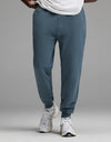 A person is shown from the waist down wearing Cozy Earth’s Men’s Dylan Jogger in blue, paired with a white shirt and white athletic sneakers, standing against a plain gray background.
