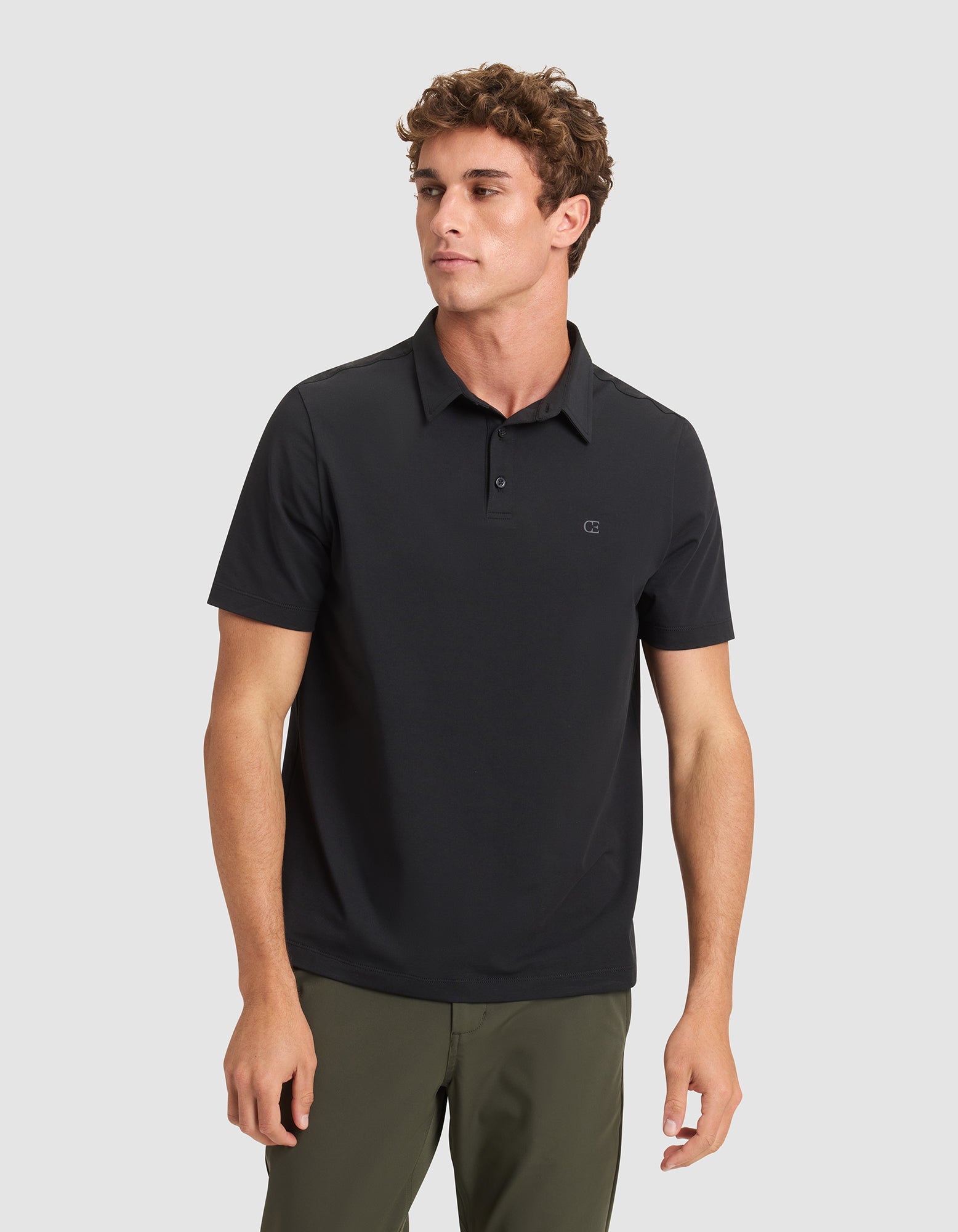 A young man with short curly hair models the Cozy Earth Men's Everyday Polo in black, paired with olive green pants, standing against a light gray background and gazing slightly to his left. 