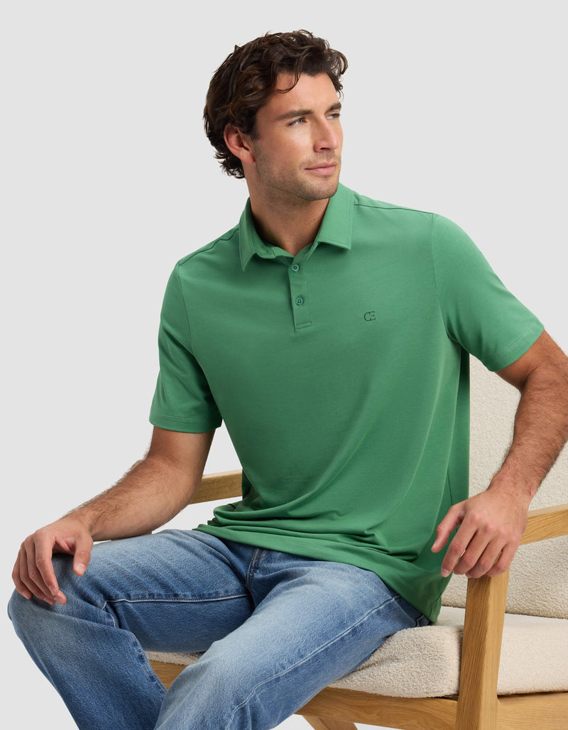 A man in a green Cozy Earth Men's Everyday Polo and blue jeans sits on a light wooden chair, gazing to the side with a relaxed expression.