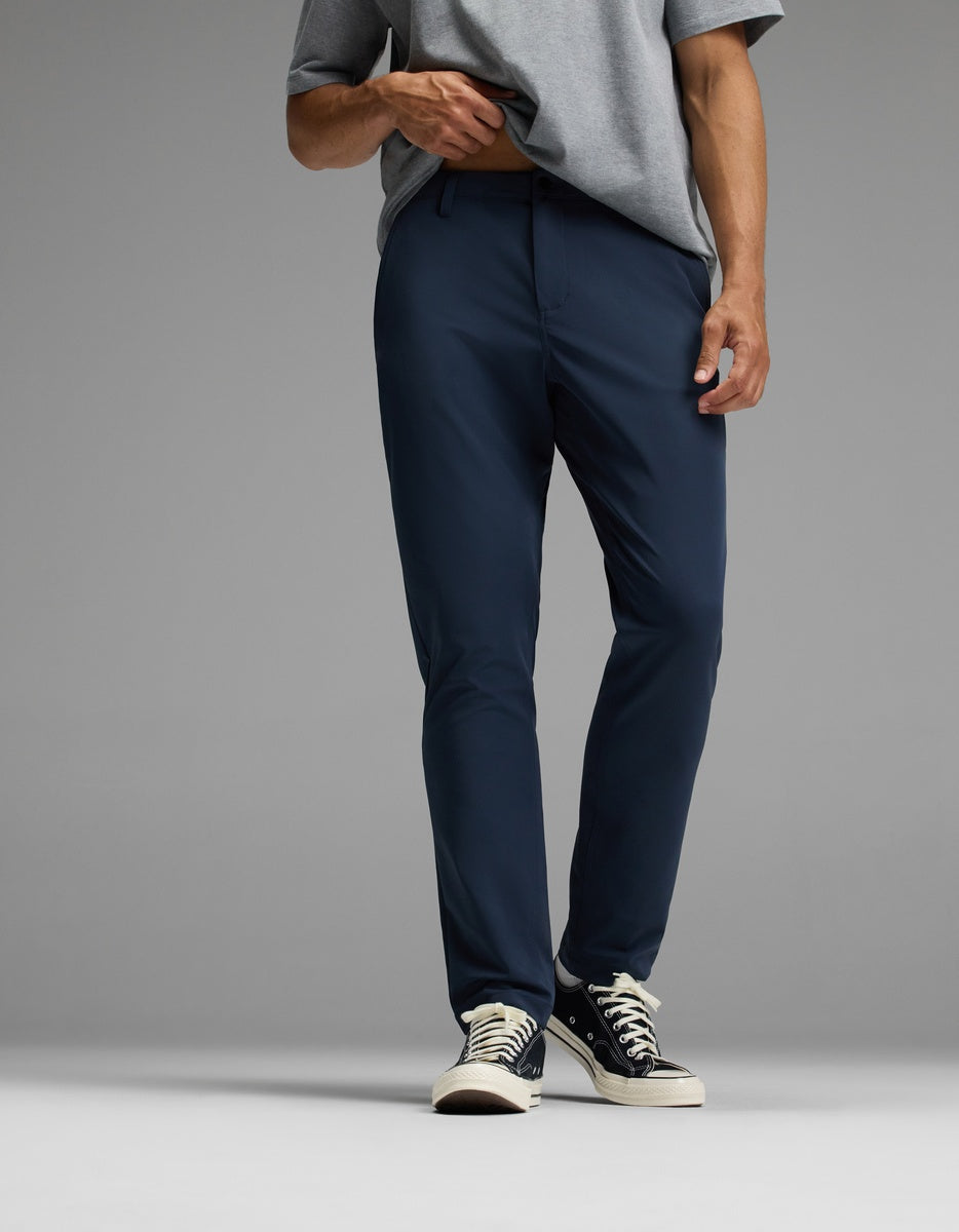 A person wearing Cozy Earth’s Men’s Everywhere Pant in navy blue, paired with a grey t-shirt and black low-top sneakers, stands against a plain grey background. Their head is not visible in the image.