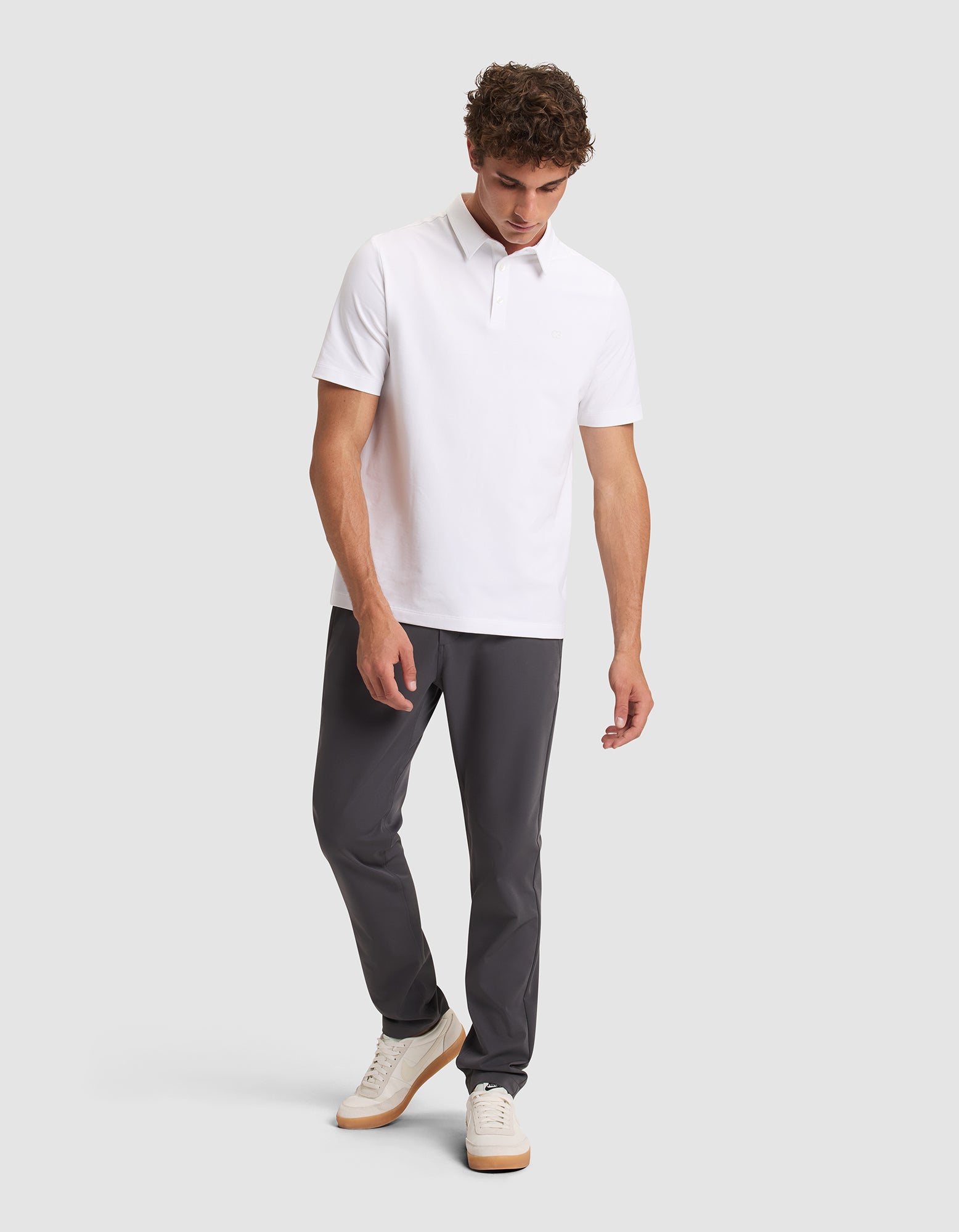 A man wearing Cozy Earth's Men's Everywhere Pant in dark gray, a white short-sleeve polo shirt, and white sneakers stands against a light gray background, looking down and slightly to the side. 