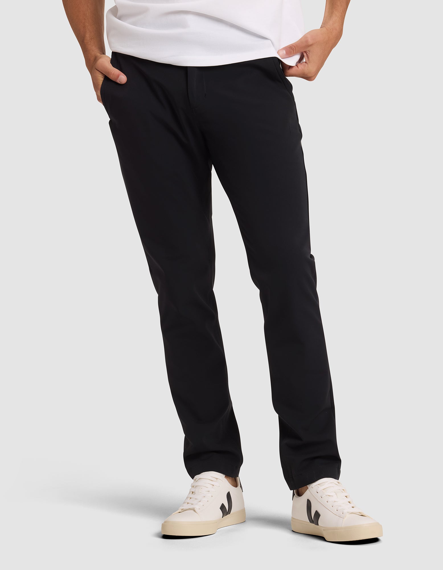 A person wearing Cozy Earth's Men's Everywhere Pant in black, a white t-shirt, and cream-colored sneakers with black detailing stands with hands in pockets against a plain light background. |Color:Jet Black