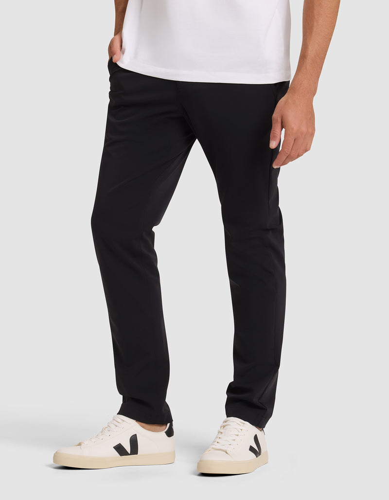 A person wearing Cozy Earth's Men's Everywhere Pant in black, a white t-shirt, and white sneakers with black detail stands against a plain light background, showing only the lower half of their body.