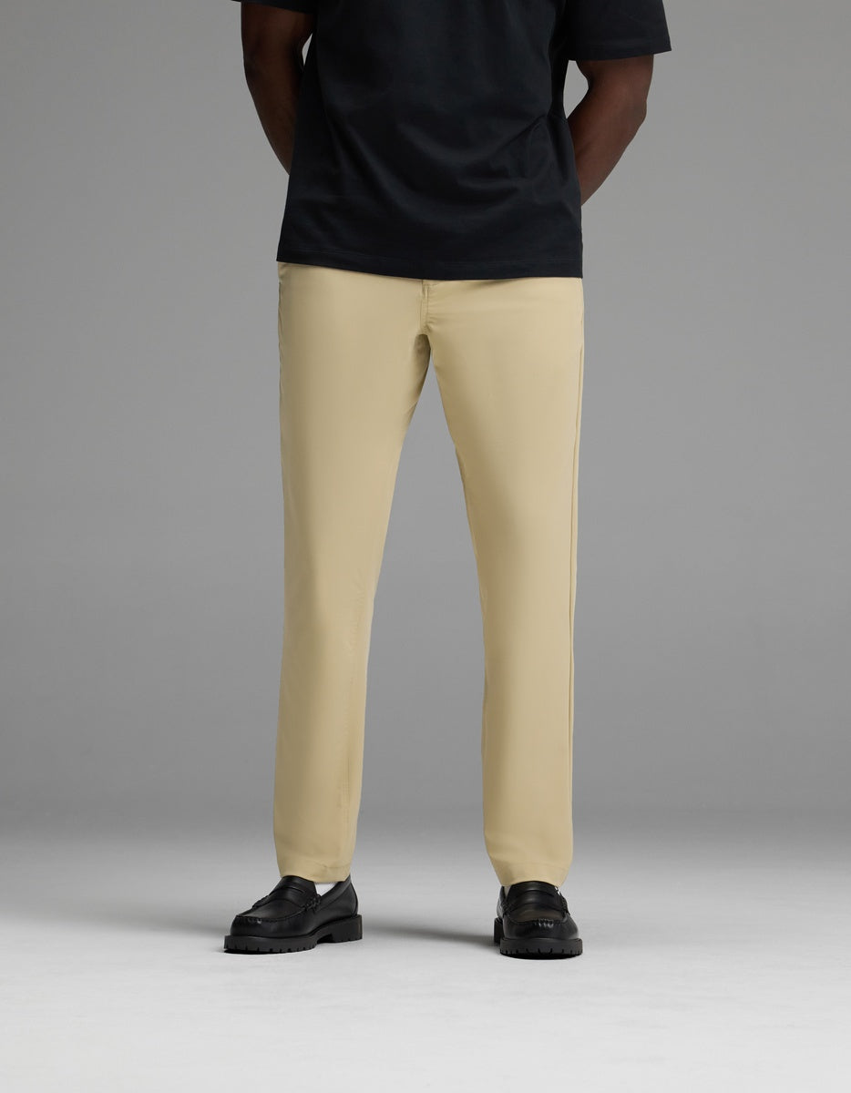 A person wearing the Cozy Earth Men’s Everywhere Pant in beige, paired with a black short-sleeve shirt and black shoes, stands against a plain gray background. Their head is not visible in the image.
