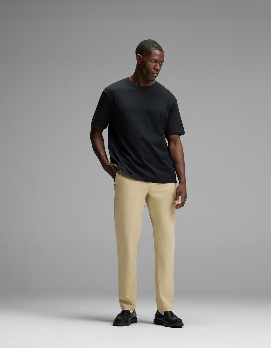 A man stands against a gray background, wearing Cozy Earth’s Men’s Everywhere Pant in beige, paired with a plain black t-shirt and black shoes. He looks down with one hand in his pocket.