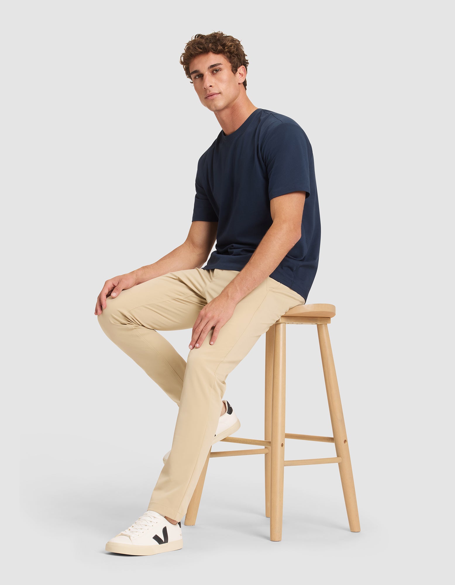 A young man sits on a wooden stool, wearing Cozy Earth's Men's Everywhere Pant in beige, a navy t-shirt, and white sneakers with a black "V" logo, against a plain light gray background. |Color:Khaki