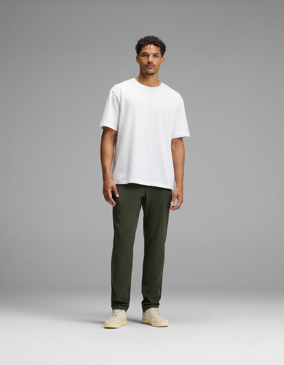 A man stands facing forward against a simple gray background, wearing olive green Men’s Everywhere Pant by Cozy Earth, a plain white t-shirt, and beige sneakers.