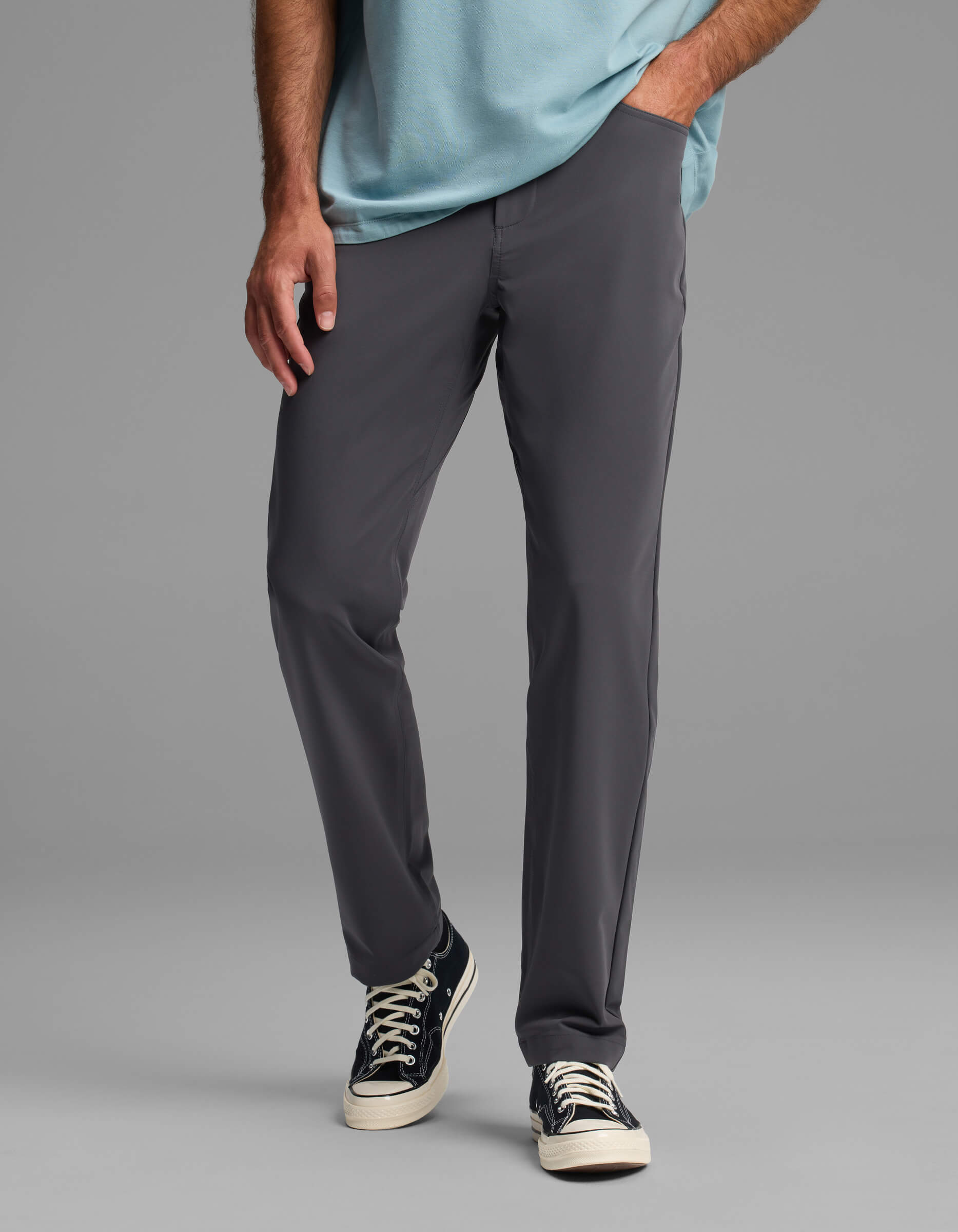 A man wears Cozy Earth's Men's Everywhere Pant - Relaxed Fit in gray, paired with a light blue shirt and black canvas sneakers with white laces, standing with one hand in his pocket against a plain gray background.