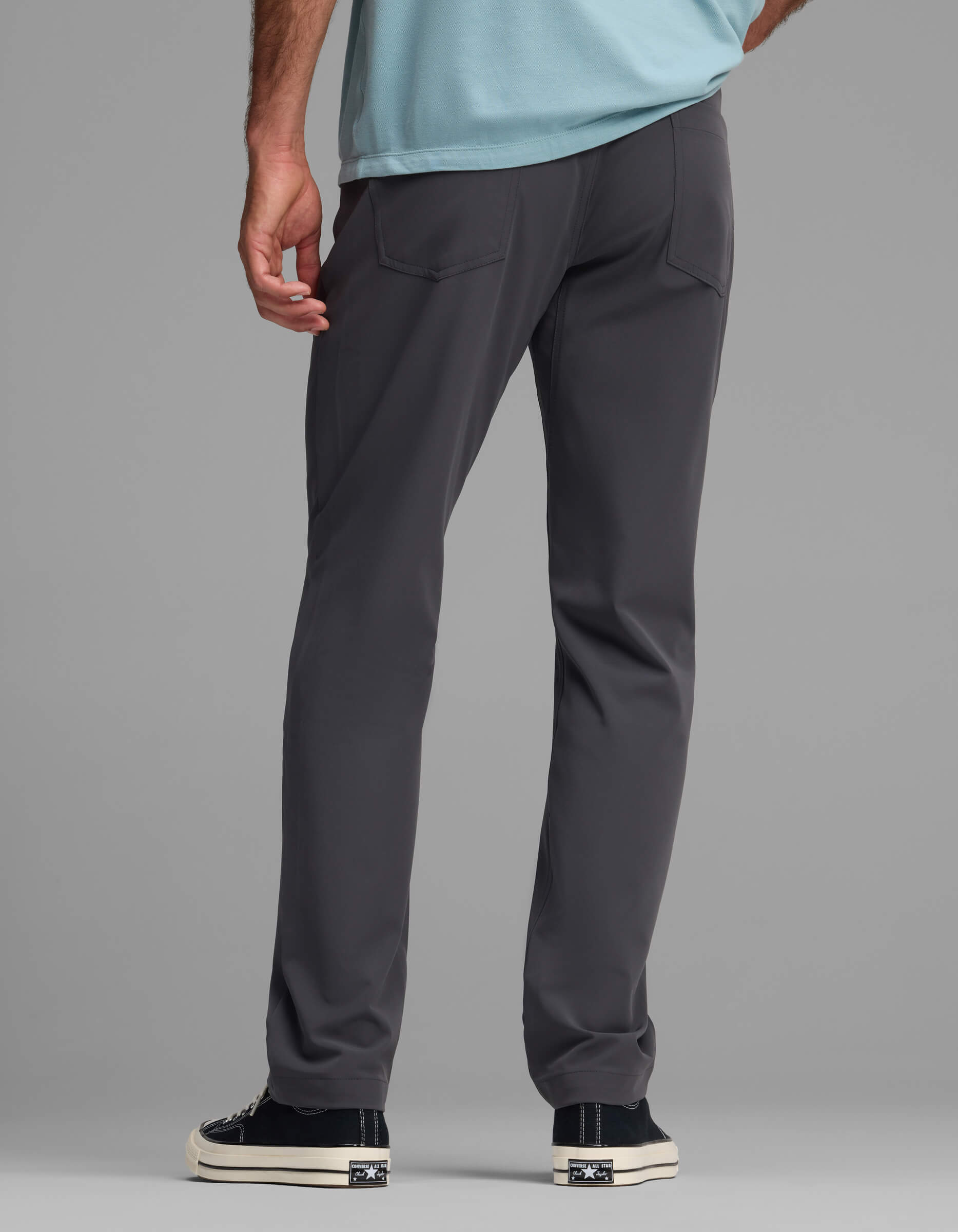 A person is seen from behind wearing the Cozy Earth Men's Everywhere Pant - Relaxed Fit in dark gray, paired with a light blue shirt and black-and-white sneakers, standing against a plain gray background.