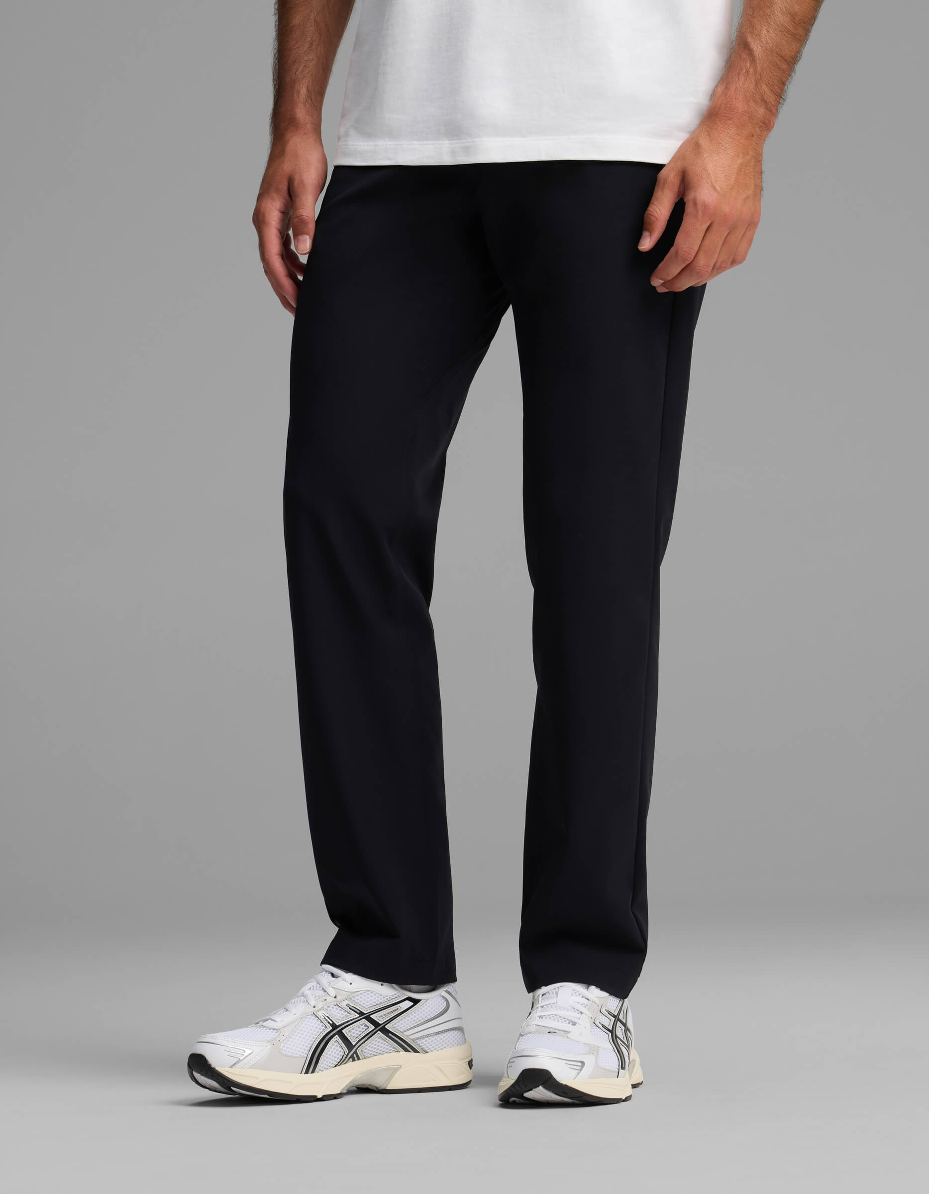 A person wears Cozy Earth's Men's Everywhere Pant - Relaxed Fit in black, paired with a white t-shirt and white sneakers with black accents. Only the lower half of the body is visible against a plain gray background.