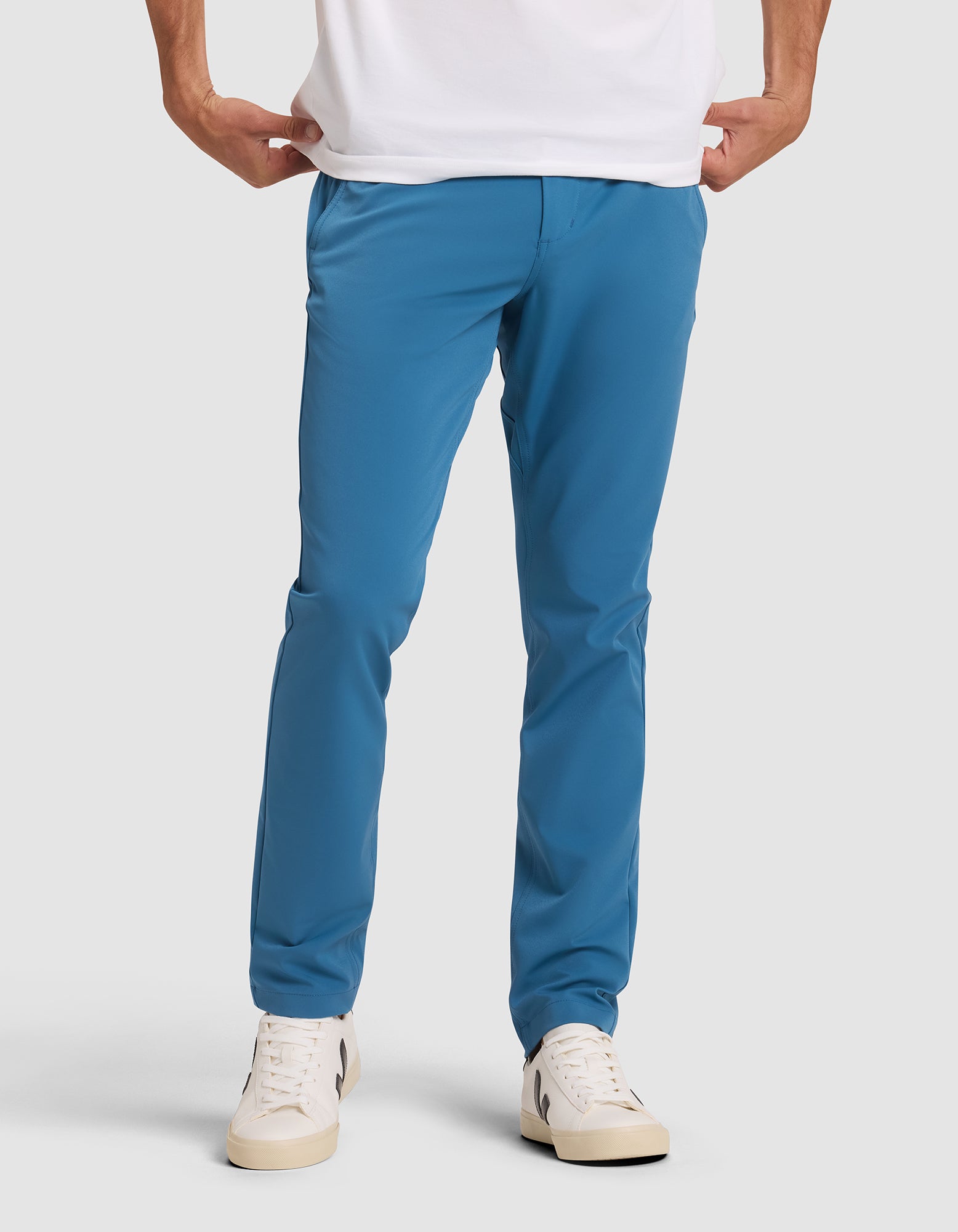 A person models Cozy Earth's Men's Everywhere Pant in bright blue with hands in pockets, paired with a white t-shirt and white sneakers, standing against a plain light gray background. The upper body is cropped from the image.  