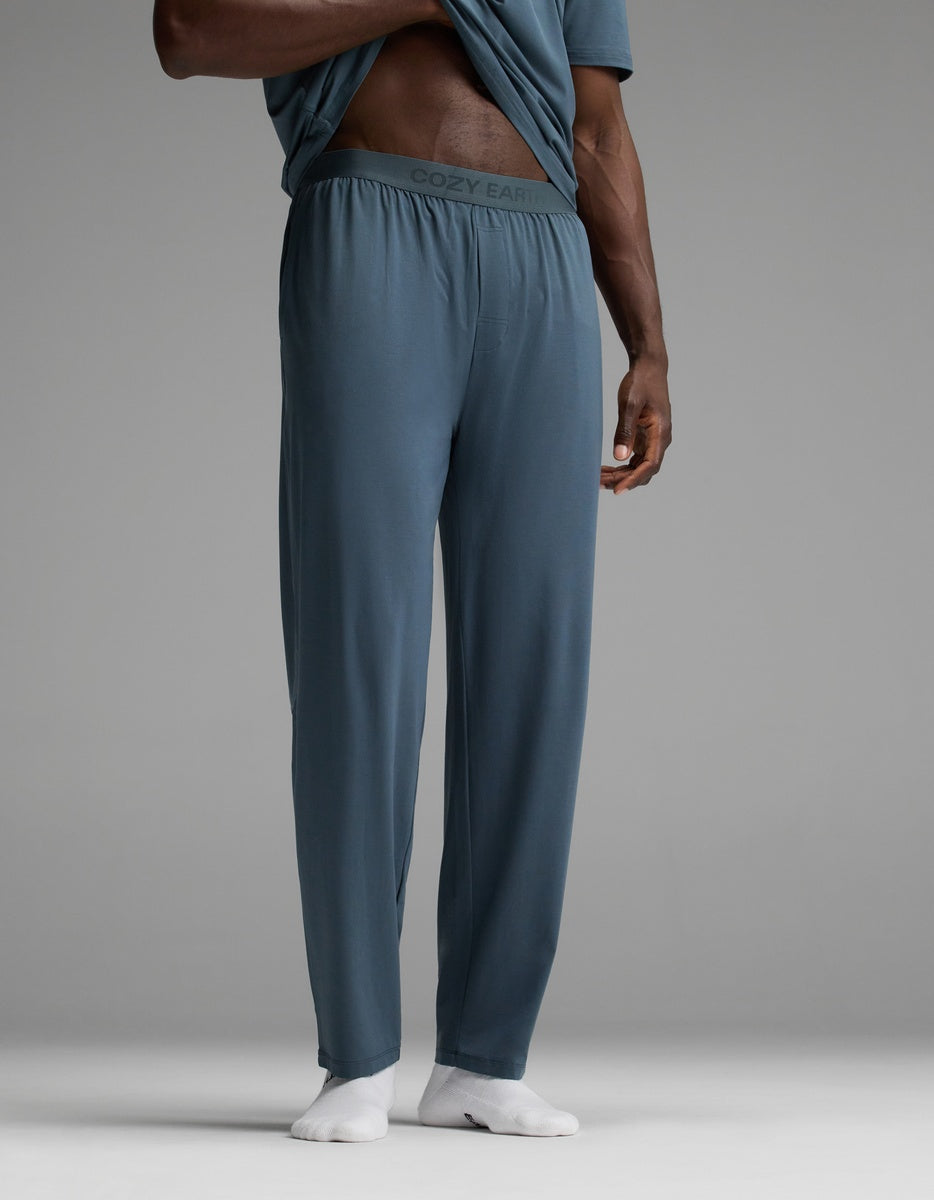A person is wearing Cozy Earth Men’s First Layer Pajama Pants in blue, featuring a waistband with “COZY EARTH” branding. They are lifting their shirt to reveal the branded waistband and are also wearing white socks. The background is plain gray.|Color:Dawn