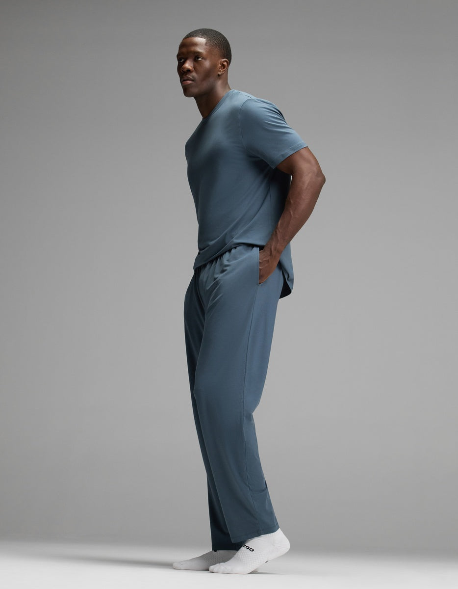 A man stands sideways against a plain gray background, wearing the Cozy Earth Men’s First Layer Pajama Pant in blue as part of a matching set with a blue t-shirt and white socks. He has his hands in his pockets.