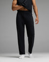A person is shown from the lower body and part of one arm, standing against a gray background. They are wearing Cozy Earth’s Men’s First Layer Pajama Pant in black, paired with a black shirt and white socks. The person lifts their shirt slightly to reveal their midsection.|Color:Jet Black