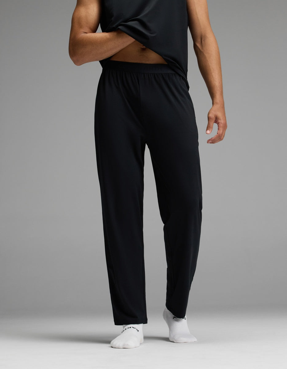 A person is shown from the lower body and part of one arm, standing against a gray background. They are wearing Cozy Earth’s Men’s First Layer Pajama Pant in black, paired with a black shirt and white socks. The person lifts their shirt slightly to reveal their midsection.