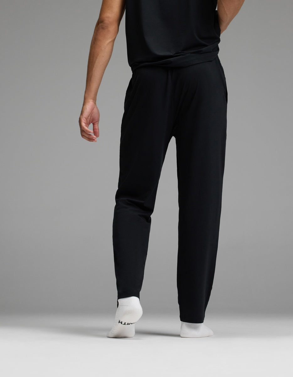 A person is seen from behind standing on a gray floor, wearing Cozy Earth Men’s First Layer Pajama Pant in black with white socks; one hand rests naturally by their side against a plain gray background.
