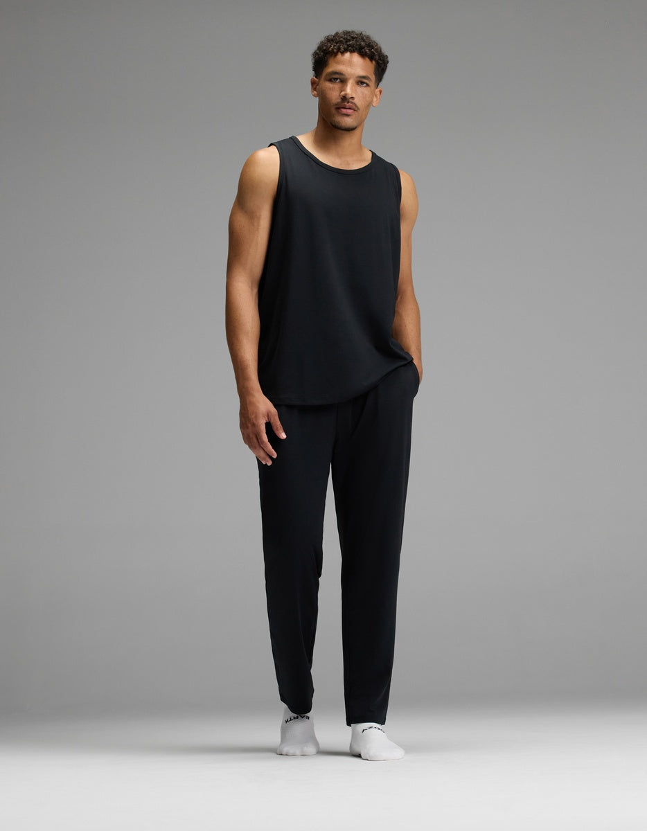 A man wearing a loose black tank top, white shoes, and Cozy Earth Men’s First Layer Pajama Pant stands against a plain gray background with one hand in his pocket.