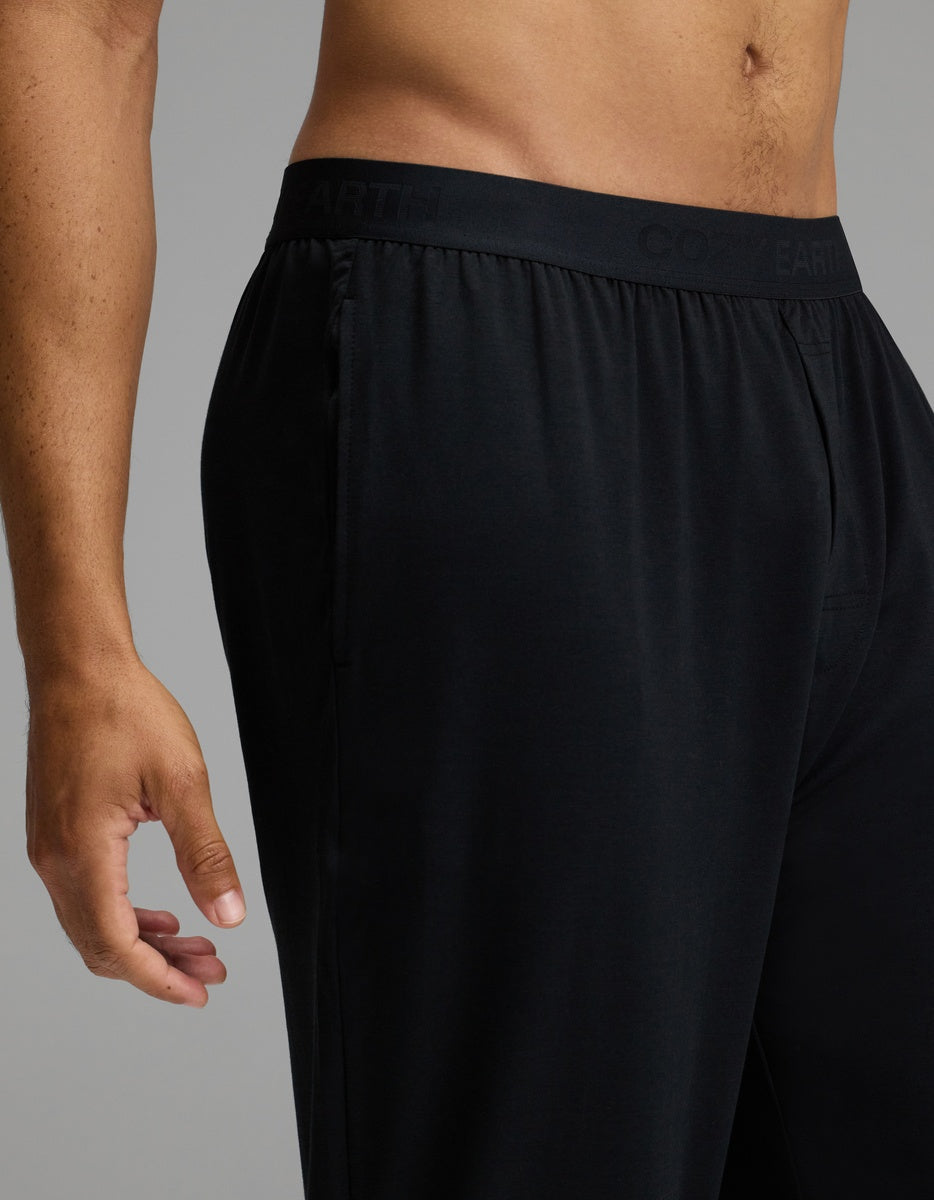 A person models the Cozy Earth Men’s First Layer Pajama Pant in black against a neutral background. The image features only the lower torso and left hand, highlighting the elastic waistband and relaxed fit of the pants.