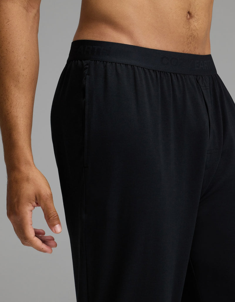 A person models the Cozy Earth Men’s First Layer Pajama Pant in black against a neutral background. The image features only the lower torso and left hand, highlighting the elastic waistband and relaxed fit of the pants.