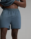 A person wears Cozy Earth’s Men’s First Layer Pajama Short in Dawn, featuring “Cozy Earth” on the waistband, lifting their shirt to reveal their torso and legs against a plain background.|Color:Dawn