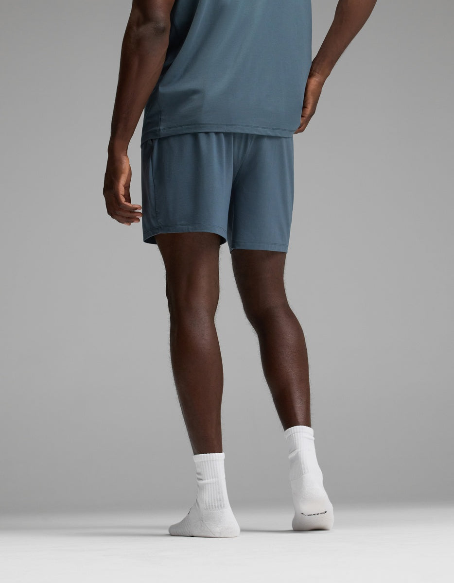 A person stands on a gray background facing away from the camera, wearing Cozy Earth Men’s First Layer Pajama Shorts in blue, a blue shirt, and white socks.