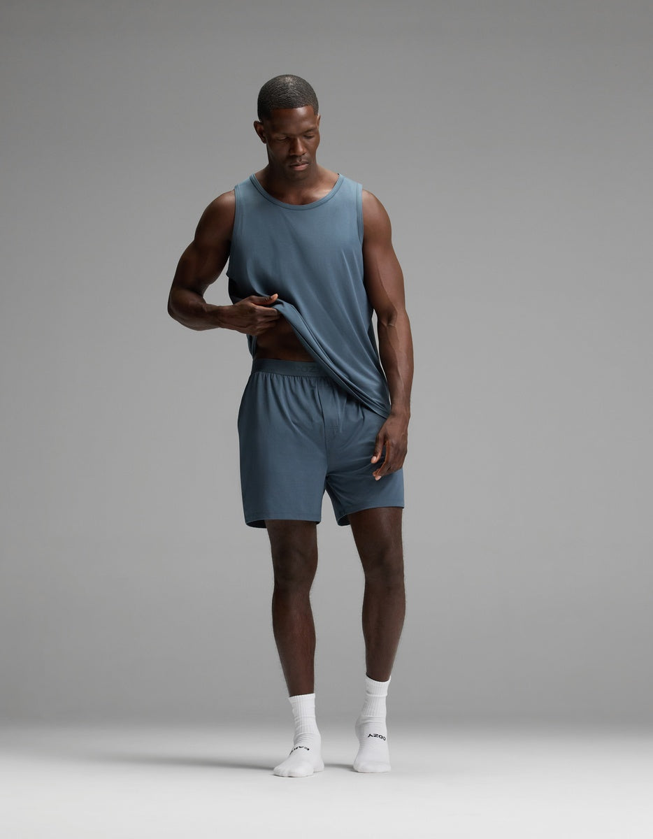 A man wearing Cozy Earth’s Men’s First Layer Pajama Short in blue stands on a gray background, paired with a matching sleeveless blue shirt and white socks, gently lifting his shirt while looking down.