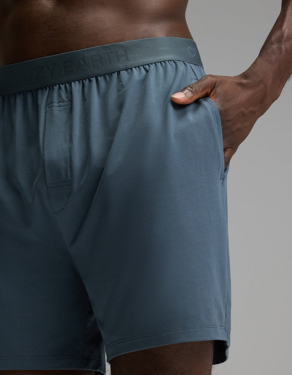 A model is shown from the lower torso to upper thighs wearing Cozy Earth Men’s First Layer Pajama Shorts in blue-gray, featuring an elastic waistband and a pocket, against a plain background.