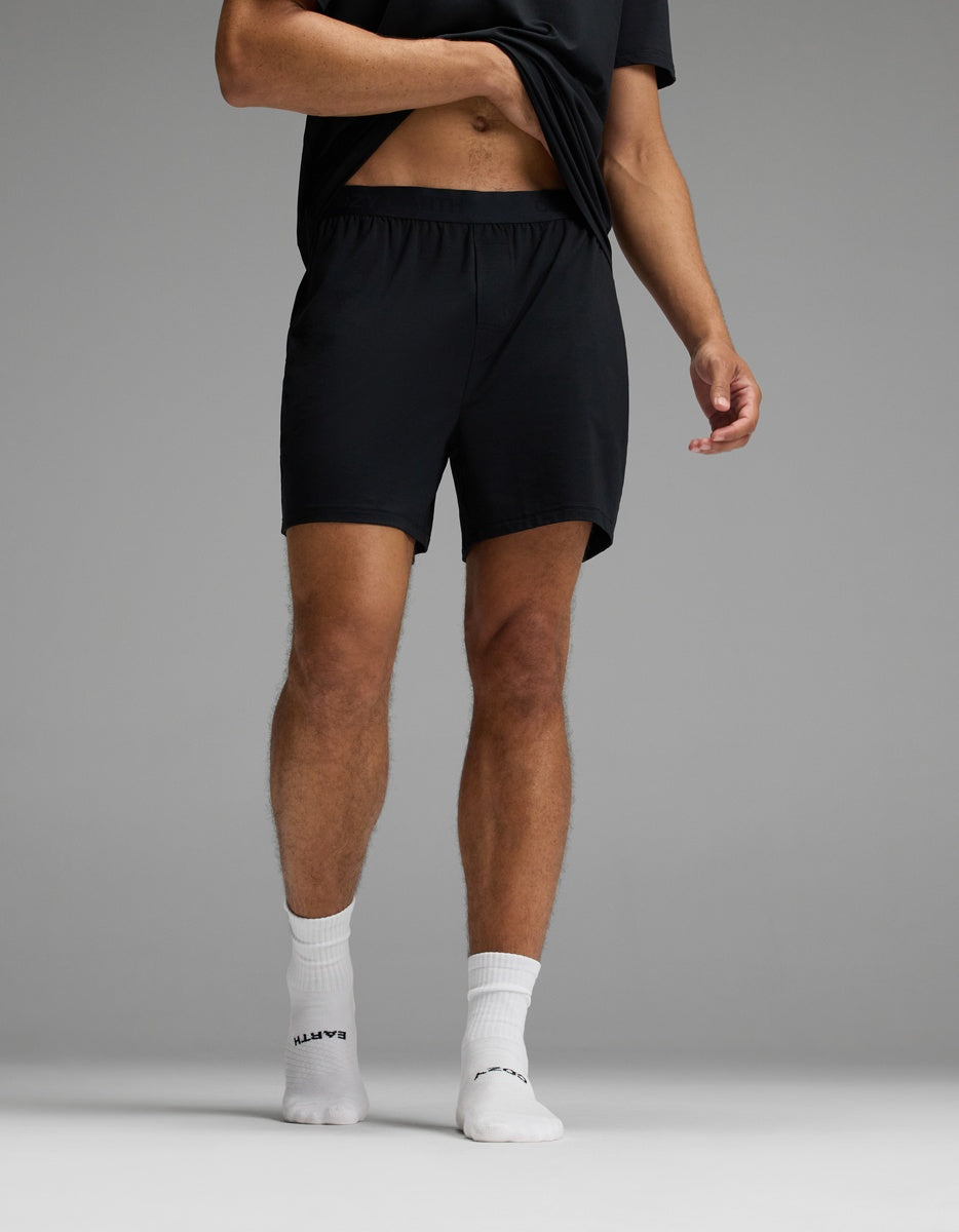 A person is standing on one foot against a plain gray background, wearing Cozy Earth Men’s First Layer Pajama Shorts paired with a black t-shirt and white socks, lifting their shirt slightly.
