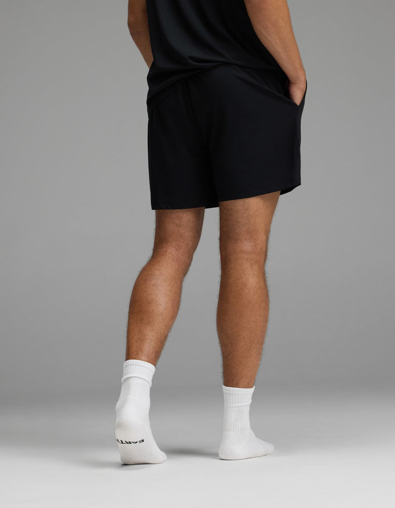 A person stands with hands in pockets, wearing the Cozy Earth Men’s First Layer Pajama Short in black, paired with a black shirt and white socks. The image displays their back and legs against a plain gray background.