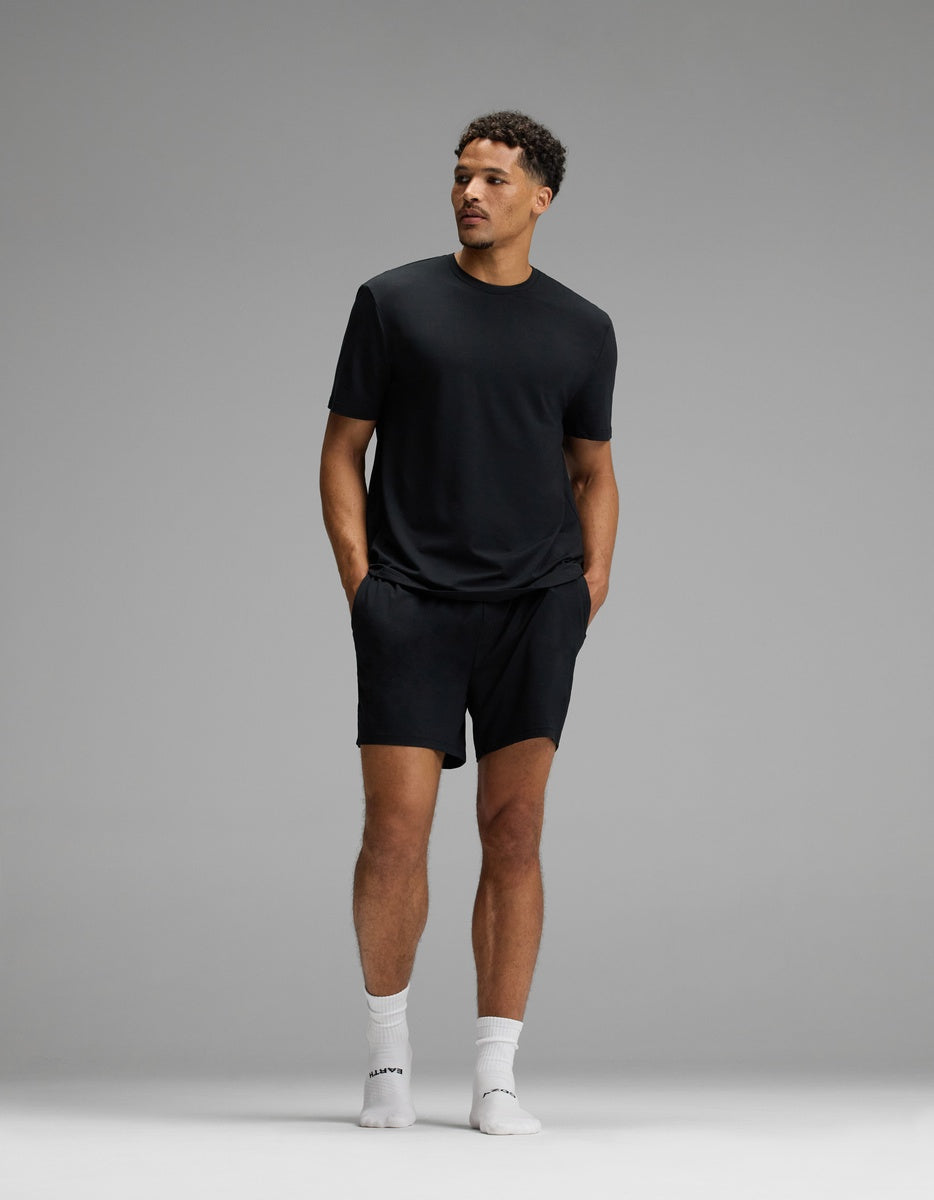 A man stands against a plain gray background, wearing Cozy Earth’s Men’s First Layer Pajama Short, paired with a black t-shirt, white crew socks, and white sneakers. His hands are in his pockets as he looks off to the side.