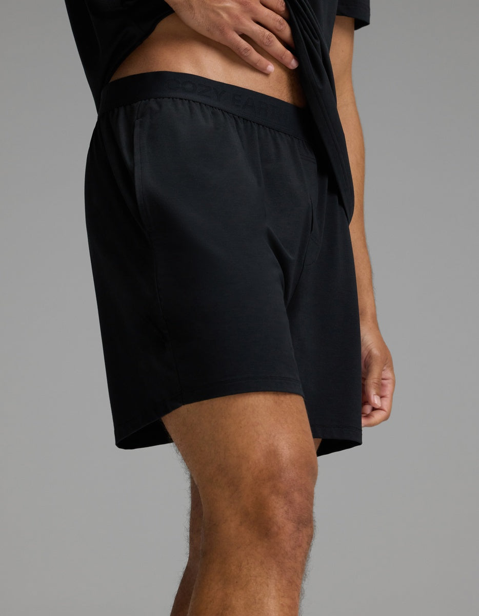 A person wearing Cozy Earth’s Men’s First Layer Pajama Short in Jet Black, paired with a matching black shirt, stands against a plain gray background with one hand resting on their waist.