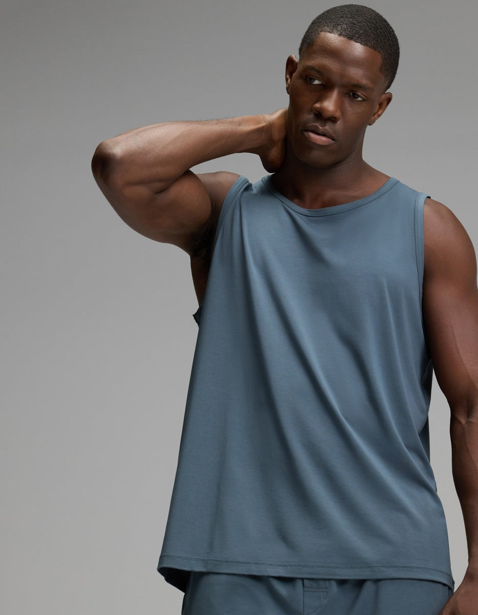 A man wearing the Cozy Earth Men’s First Layer Pajama Tank and matching shorts in Dawn stands against a plain gray background, with one hand resting behind his neck.