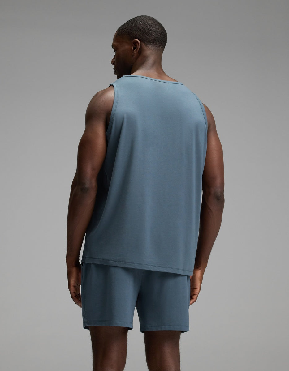 A man stands facing away from the camera, wearing the Cozy Earth Men’s First Layer Pajama Tank in blue, paired with matching blue shorts, against a plain gray background.