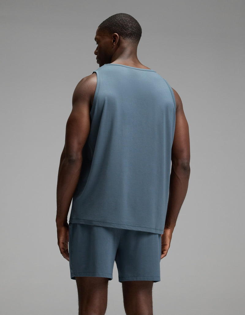 A man stands facing away from the camera, wearing the Cozy Earth Men’s First Layer Pajama Tank in blue, paired with matching blue shorts, against a plain gray background.