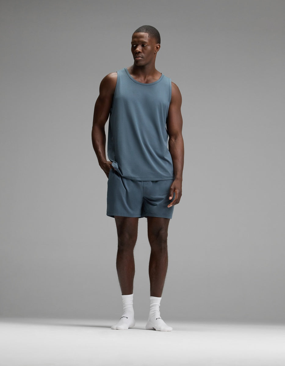 A man wearing the Cozy Earth Men’s First Layer Pajama Tank in blue, with matching shorts and white socks, stands against a plain gray background, looking to his left with one hand in his pocket.