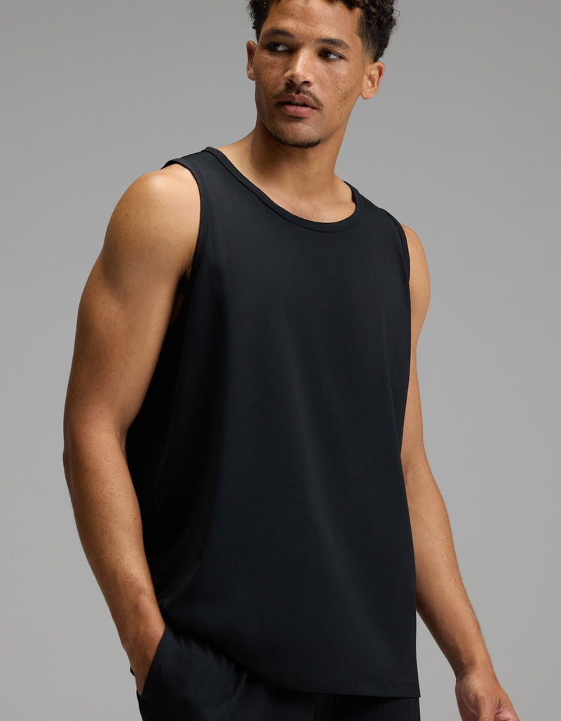 A man wears the Cozy Earth Men’s First Layer Pajama Tank in Jet Black, paired with matching black pants, as he stands against a plain gray background and looks to his left with a neutral expression.