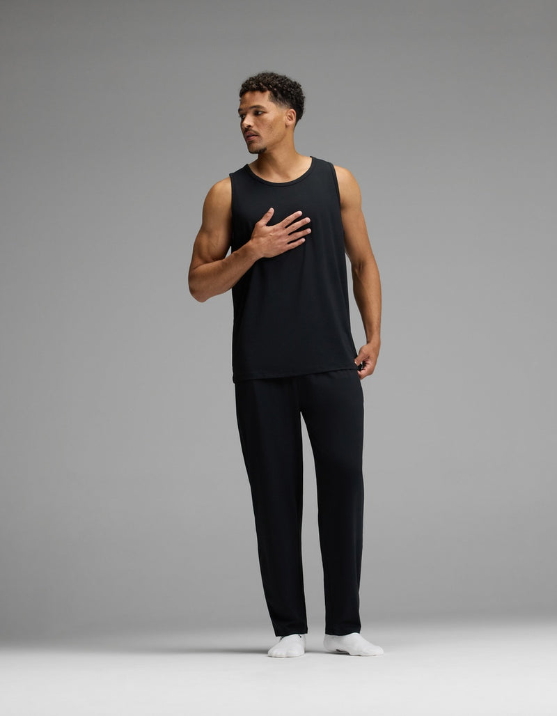 A man wearing the Cozy Earth Men’s First Layer Pajama Tank in black and matching black pants stands with one hand on his chest against a plain gray background, paired with white socks.
