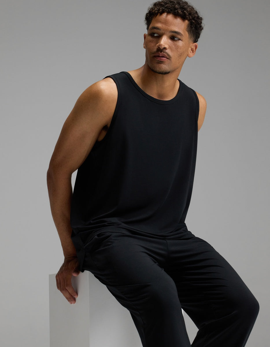 A man wearing the Cozy Earth Men’s First Layer Pajama Tank in black and matching black pants sits on a white block, gazing to the side against a plain gray background.