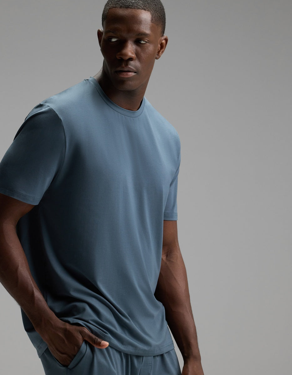 A man wearing the Cozy Earth Men’s First Layer Pajama Short Sleeve Tee in Dawn, along with matching pants, stands with one hand in his pocket and looks to his left against a plain gray background.
