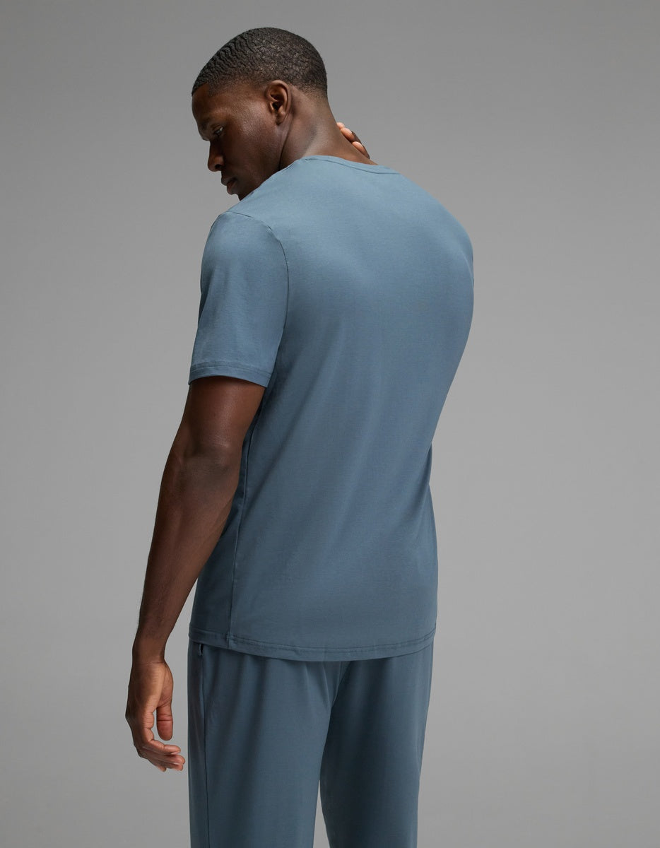 A man models the Cozy Earth Men’s First Layer Pajama Short Sleeve Tee in blue with matching pants, standing against a plain gray background, his head turned slightly to the side.