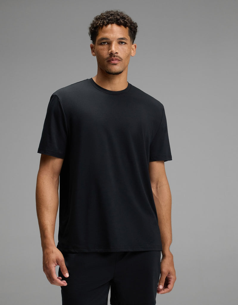 A man with short curly hair wears the Cozy Earth Men’s First Layer Pajama Short Sleeve Tee in Jet Black along with black pants, standing against a plain gray background. He faces forward with a neutral expression.