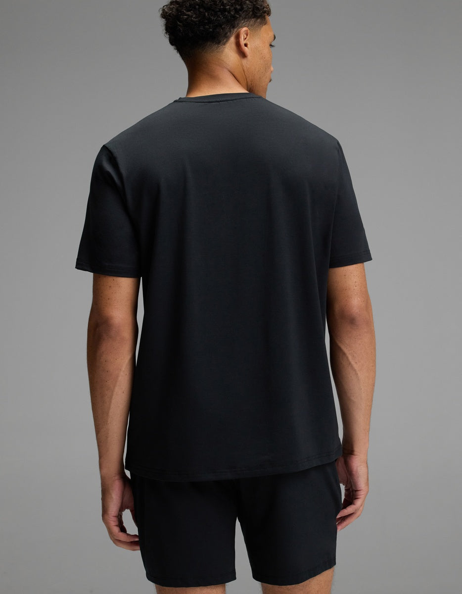 A man stands facing away from the camera, wearing the Cozy Earth Men’s First Layer Pajama Short Sleeve Tee in black and matching black shorts against a plain gray background.