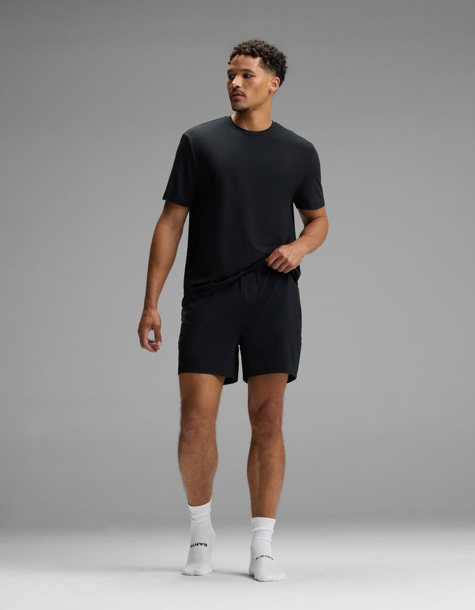 A man stands in a studio against a plain gray background, wearing the Cozy Earth Men’s First Layer Pajama Short Sleeve Tee in black, paired with black shorts and white socks. He looks to his right with one hand by his side and the other touching his shorts.