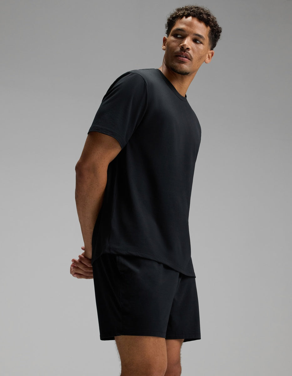 A man wearing a Cozy Earth Men’s First Layer Pajama Short Sleeve Tee and black shorts stands against a plain gray background, looking slightly to the side with his hands behind his back.